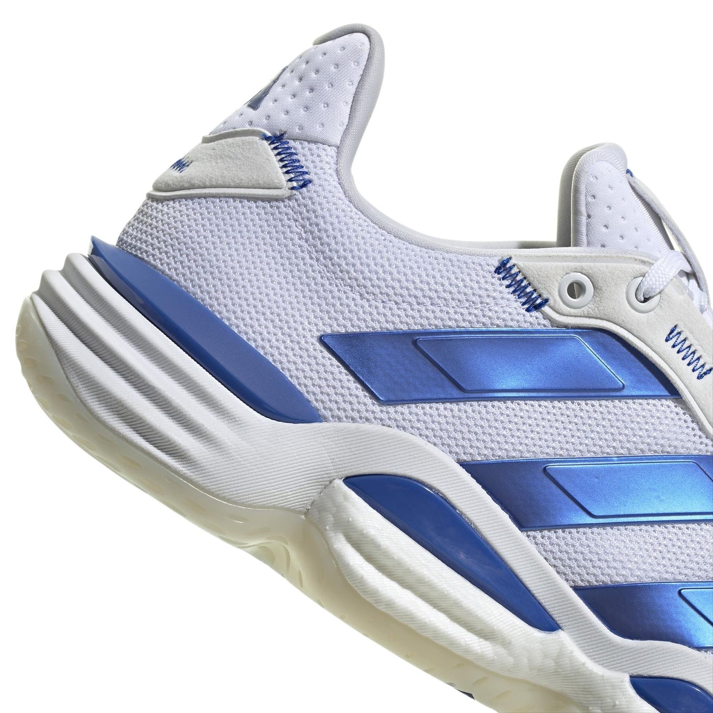 Close-up of the adidas Stabil 16 Men's Indoor Court Shoes in white/blue with blue metallic stripes, textured fabric upper, padded ankle, BOOST midsole, and chunky sole. An inscription is visible on the side.