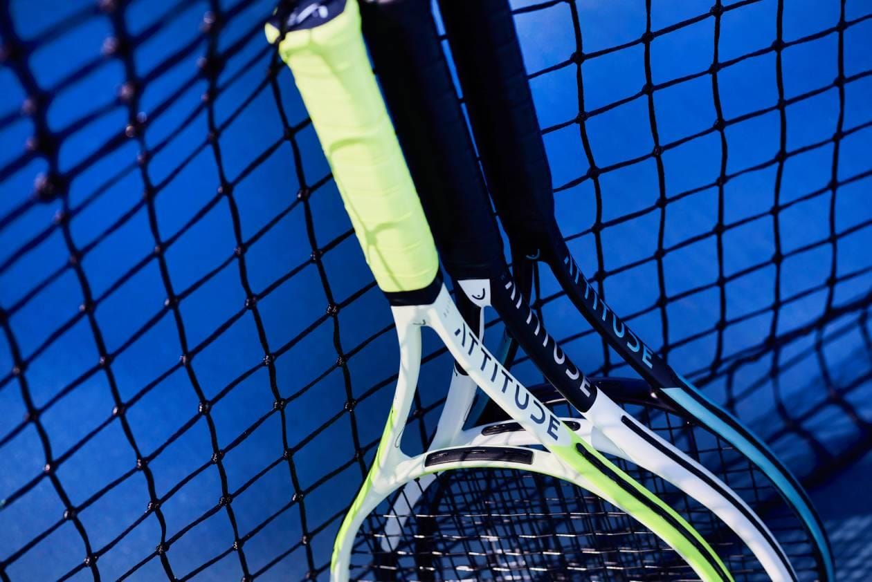 Three lightweight HEAD MX Attitude Elite Tennis Rackets by HEAD, featuring green and black handles, lean against a black net on a blue tennis court, seen from above.