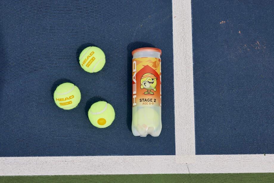 Three orange HEAD 3B Stage 2 Tennis Balls sit by a canister on a blue court near the white line, ideal for kids ages 6–9 to learn and play with quality HEAD Stage 2 ORANGE balls.