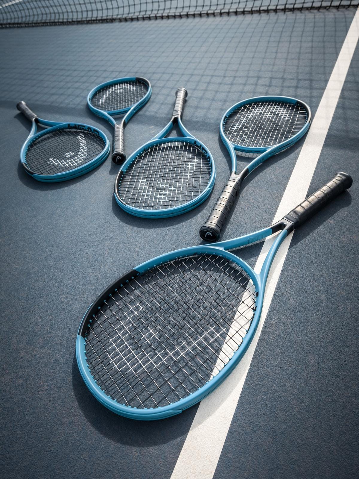 Five HEAD Boom MP L 2026 tennis rackets in blue and black, known for their effortless power, are arranged on a blue tennis court near the white lines with a tennis net visible in the background.