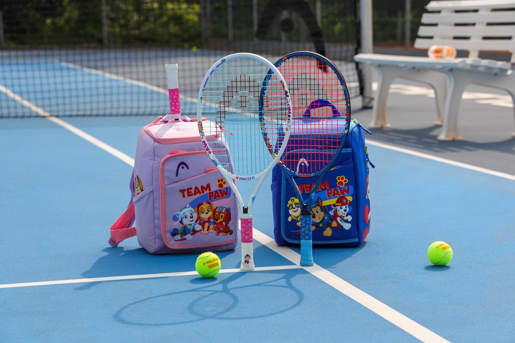 Two kids' tennis backpacks, including the HEAD x Paw Patrol Kids Tennis Backpack in purple, each with a junior tennis racket bag and water bottle, rest on a blue court, with tennis balls nearby and a white bench in the background.