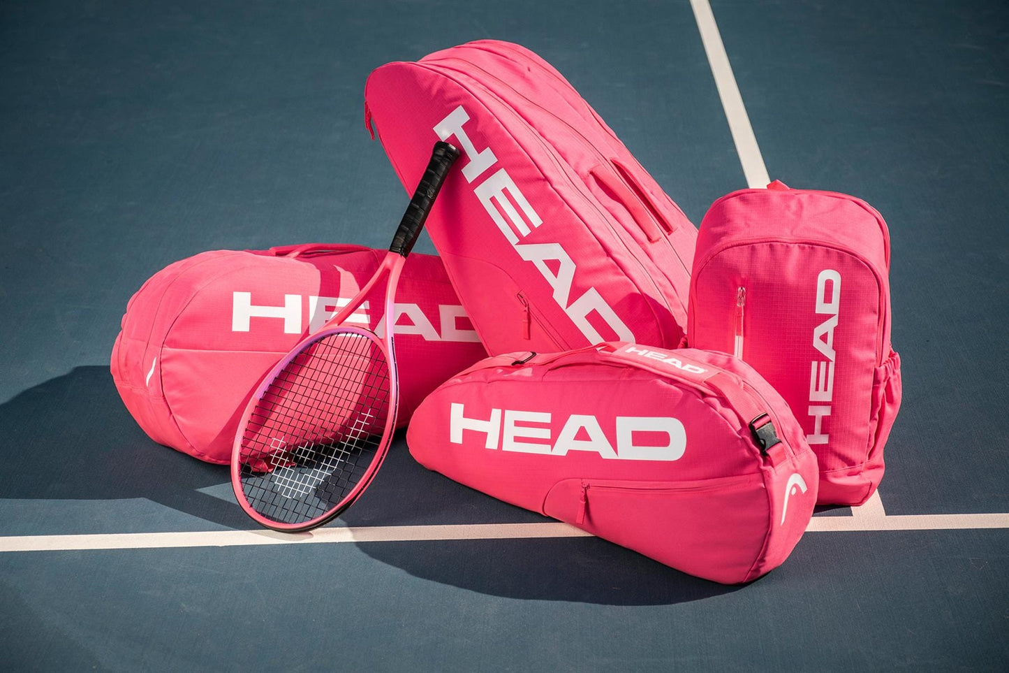 Four bright pink HEAD Base 6 Racket Tennis Bag M – Pink by HEAD, along with a matching pink tennis racket, are arranged on a blue tennis court over crisp white lines.