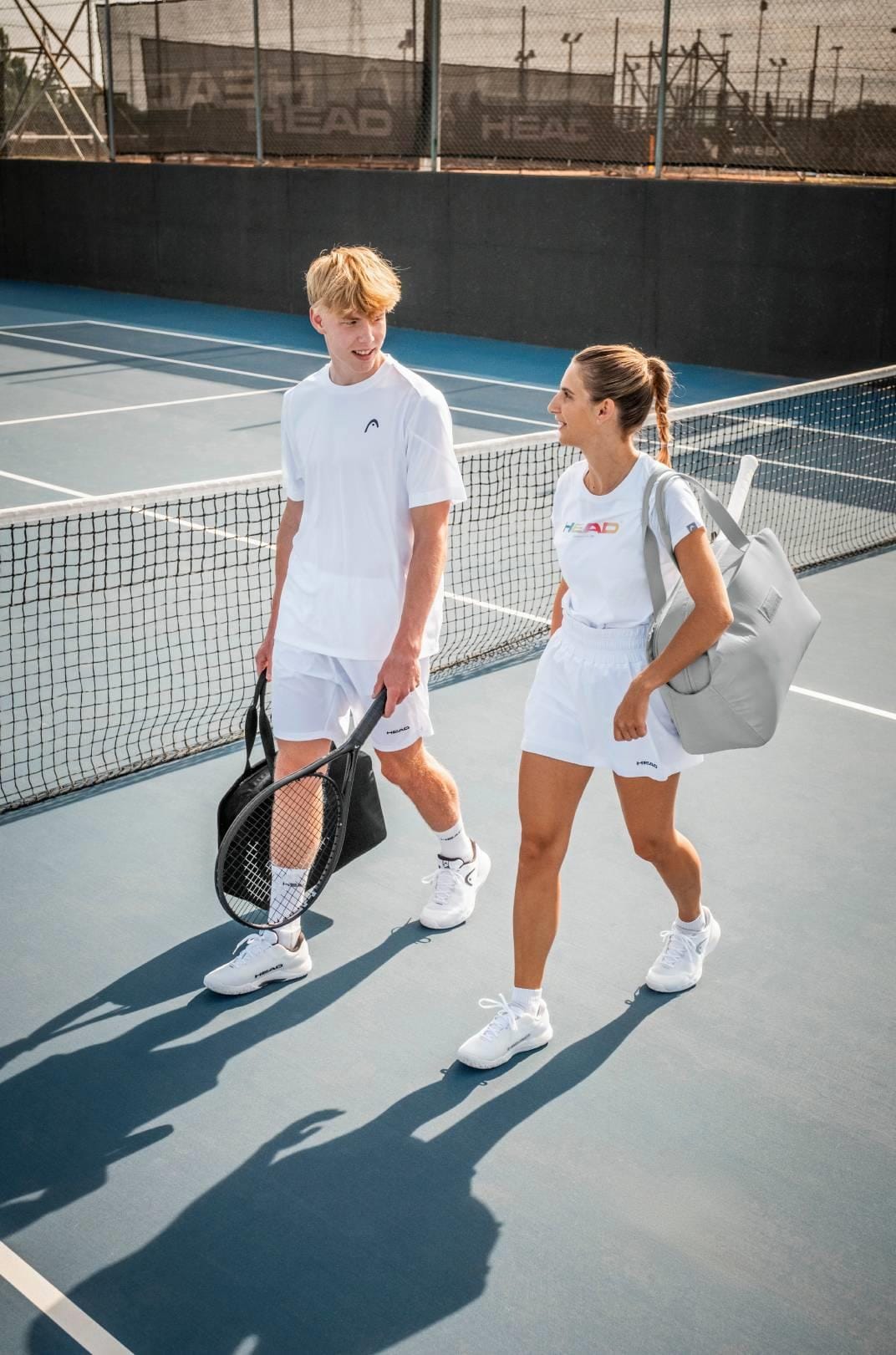 A young man and woman, both in white athletic wear, walk and chat on an outdoor court carrying HEAD PWR 110 Tennis Rackets by HEAD, looking relaxed and cheerful as they enjoy their powerful tennis gear.