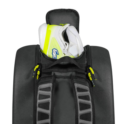 A pair of white athletic shoes with yellow accents are stored in the open top compartment of the spacious Tecnifibre Reform L Tennis Racket Bag - Black, featuring black, gray, and yellow zipper details.