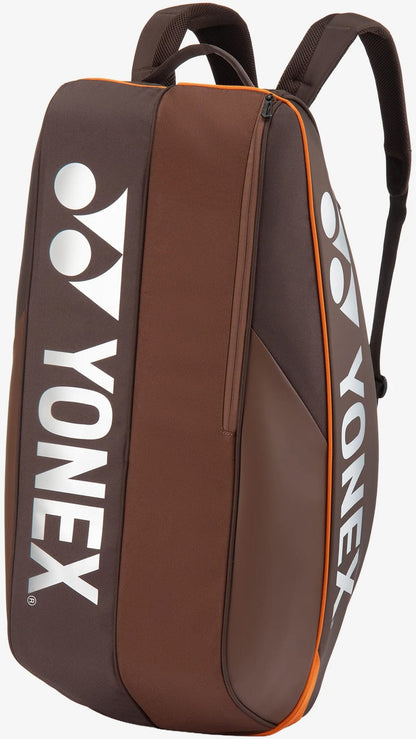 The Yonex 92426EX 6 Racket Pro Tennis Bag in Espresso Brown features orange trim, white Yonex logos, a convenient side zipper pocket, and two adjustable shoulder straps. Designed as a versatile tennis bag by Yonex, it offers enough space to carry up to six rackets and is pictured standing upright against a light background.