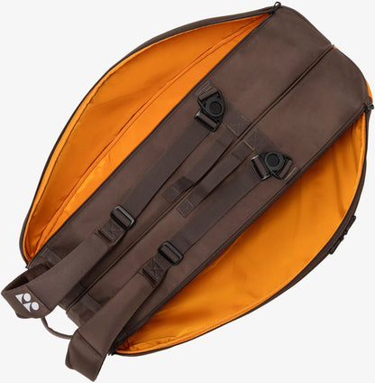 The Yonex 92426EX 6 Racket Pro Tennis Bag in Espresso Brown lies open, revealing its vibrant orange interior lining. Reminiscent of a classic 6 racket bag, this Yonex bag features two padded shoulder straps with black plastic buckles and exterior adjustment points.