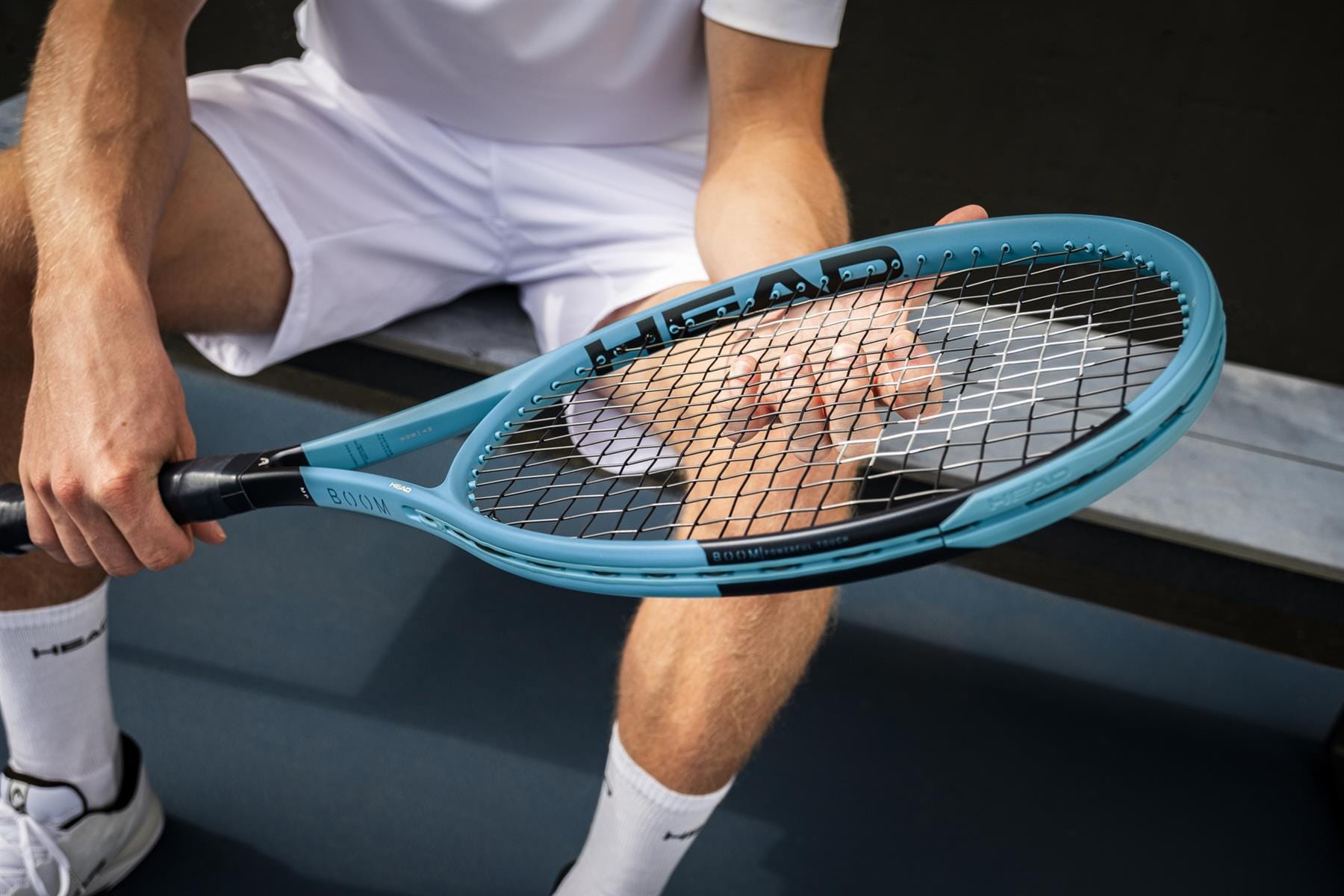 Dressed in white sportswear, a person sits on a bench at an outdoor tennis court, examining the strings of their HEAD Boom MP 2026 Tennis Racket, emphasizing the explosive power this HEAD racket delivers.