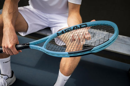 Dressed in white sportswear, a person sits on a bench at an outdoor tennis court, examining the strings of their HEAD Boom MP 2026 Tennis Racket, emphasizing the explosive power this HEAD racket delivers.