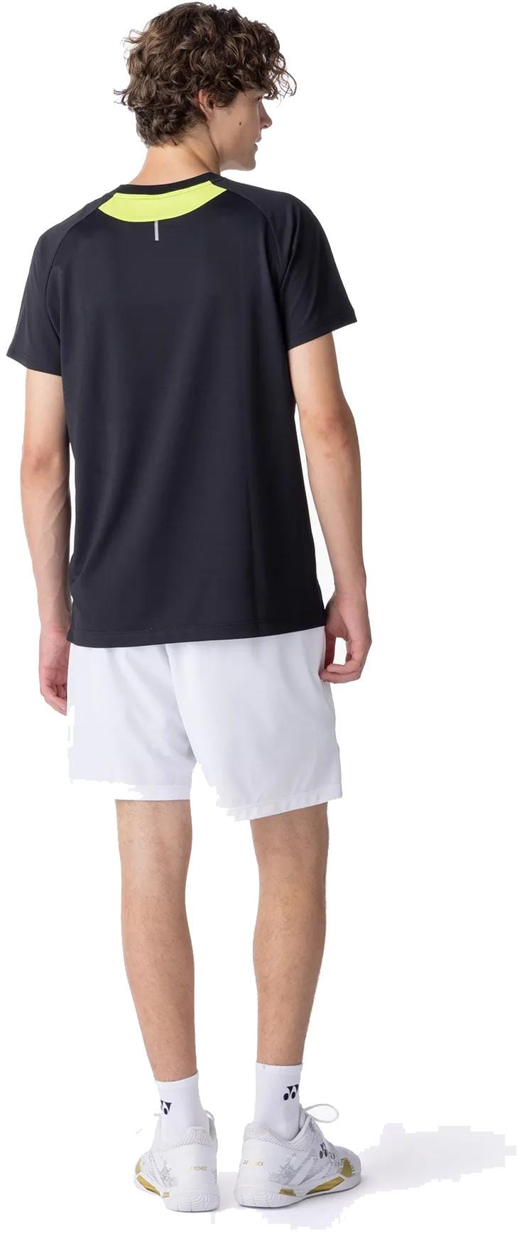 A young man with curly hair, seen from behind, wears the Yonex 10711YX Crew Neck Men's Tennis T-Shirt in black, paired with white athletic shorts, socks, and sneakers against a white background.