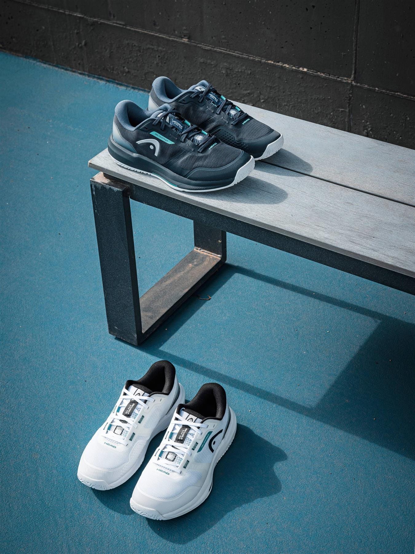 Two pairs of HEAD sneakers, including HEAD Revolt Evo 5.0 Men's Tennis Shoes in White/Black, are shown on a gray bench and the blue floor against a dark wall. The black pair sits on the bench; the white/black tennis shoes rest on the ground.