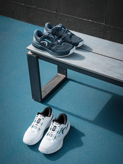 Two pairs of HEAD sneakers, including HEAD Revolt Evo 5.0 Men's Tennis Shoes in White/Black, are shown on a gray bench and the blue floor against a dark wall. The black pair sits on the bench; the white/black tennis shoes rest on the ground.