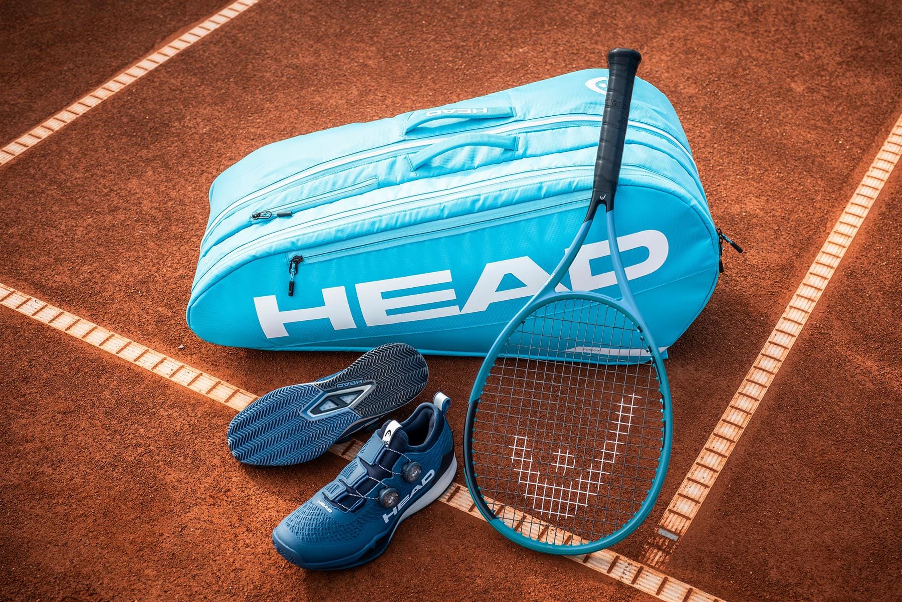 A HEAD Tour 9 Racket Tennis Bag L - Blue, a matching tennis racquet, blue tennis shoes, and shoe soles by HEAD are displayed on a red clay court.