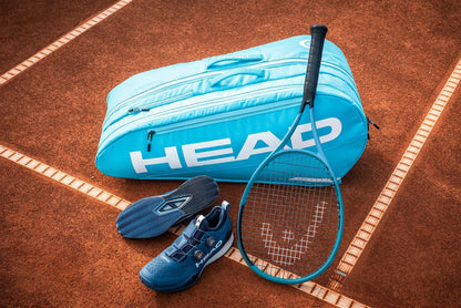 A HEAD Tour 9 Racket Tennis Bag L - Blue, a matching tennis racquet, blue tennis shoes, and shoe soles by HEAD are displayed on a red clay court.