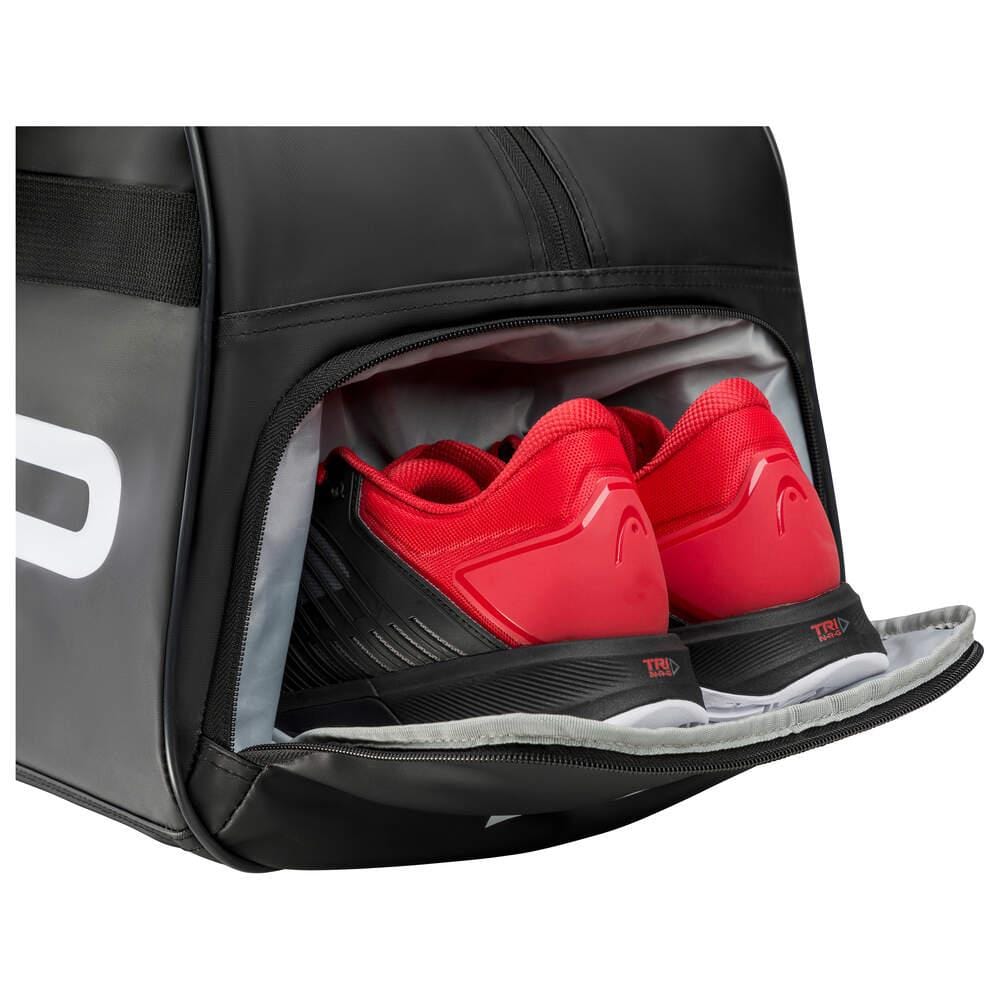 A pair of red and black sports shoes pokes out from the shoe compartment of a HEAD Tour Tennis Court Bag. Featuring a sleek black and white exterior with a gray-lined interior, the HEAD Tour Tennis Court Bag 40L effortlessly sits on a white background, highlighting its practicality and adjustable handle straps for easy carrying.