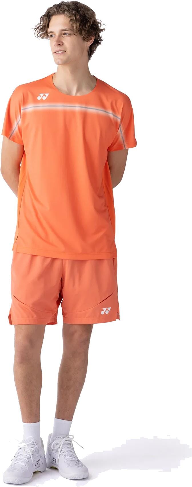 A young man stands with hands behind his back, wearing the Yonex 10711YX Crew Neck Men's Tennis T-Shirt in Cyber Orange—premium sportswear with white logo and stripes—paired with matching shorts, white socks, sneakers, and gazing slightly left.