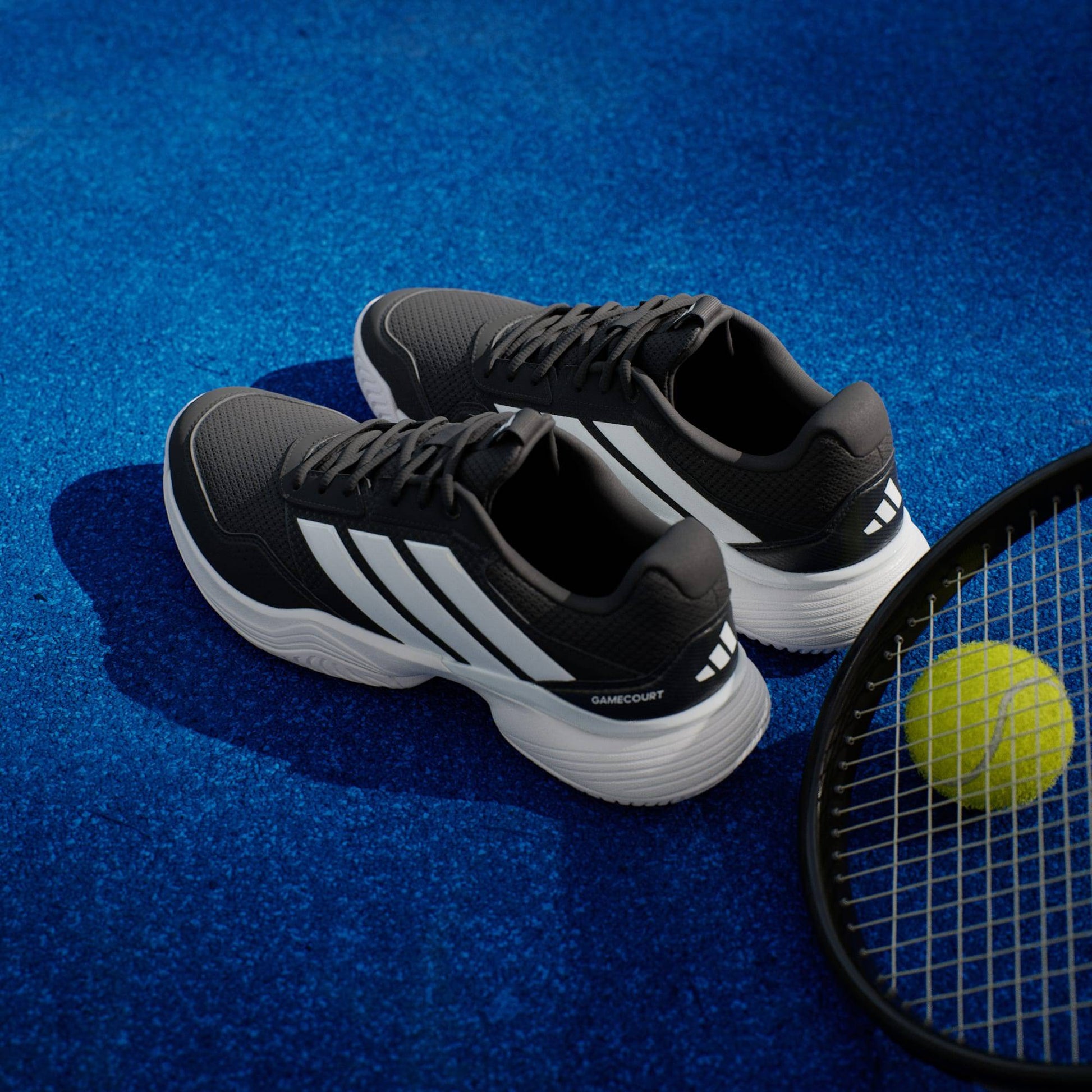 A pair of adidas Gamecourt 3 Mens Tennis Shoes - Core Black rests on a blue tennis court beside a racket and yellow ball, highlighting these durable black-and-white adidas men’s tennis shoes.