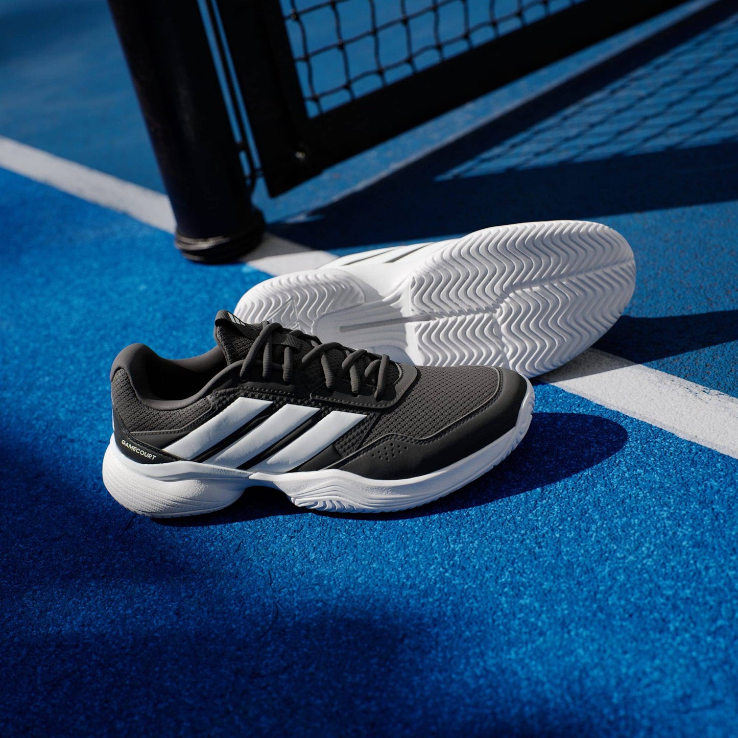A pair of adidas Gamecourt 3 Mens Tennis Shoes - Core Black is set on a blue tennis court near the white line and black net post, with sunlight highlighting these lightweight adidas tennis shoes.