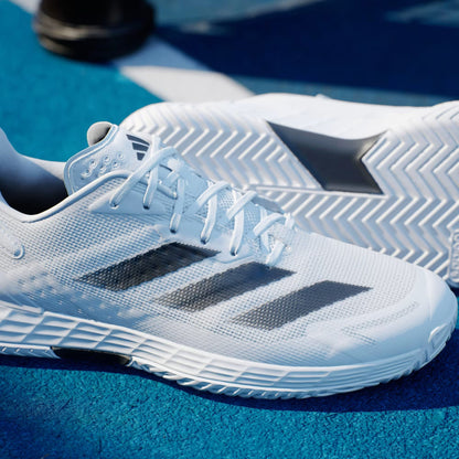 A close-up of the adidas Defiant Speed 2 Men's Tennis Shoes in Cloud White/Core Black/Grey, featuring a dark three-stripe design on mesh uppers with textured soles, resting on a blue court—perfect for speed, running, or training.