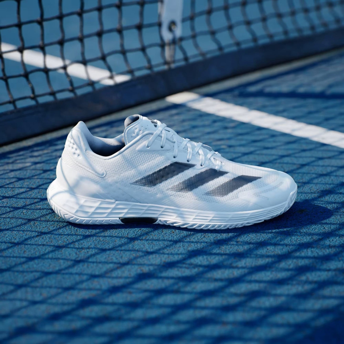 The adidas Defiant Speed 2 Men's Tennis Shoes in Cloud White, Core Black, and Grey stand on a blue court near the net, their sporty design and lightweight build emphasized by shadows cast across the surface.