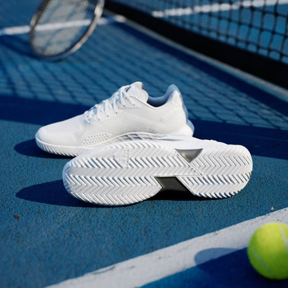 A pair of adidas Defiant Speed 2 Mens Tennis Shoes in Cloud White/Core Black/Grey are on a blue tennis court by the net, with a yellow tennis ball and racket nearby as sunlight creates shadows across the court.