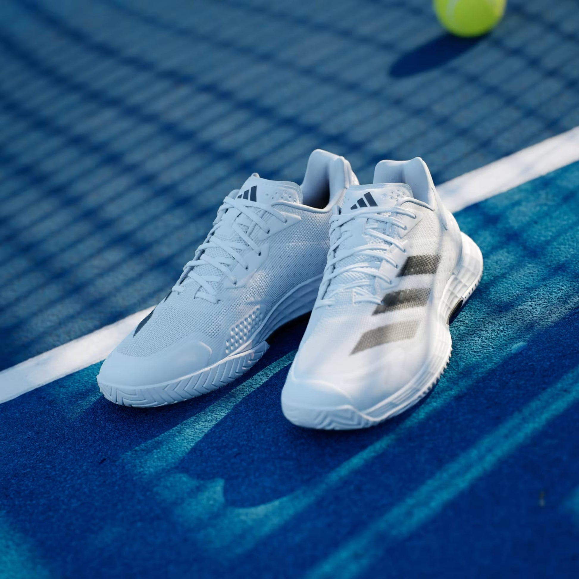 A pair of adidas Defiant Speed 2 Men's Tennis Shoes in Cloud White, Core Black, and Grey sit on a blue tennis court near the white line, with a yellow tennis ball and net visible in the background.