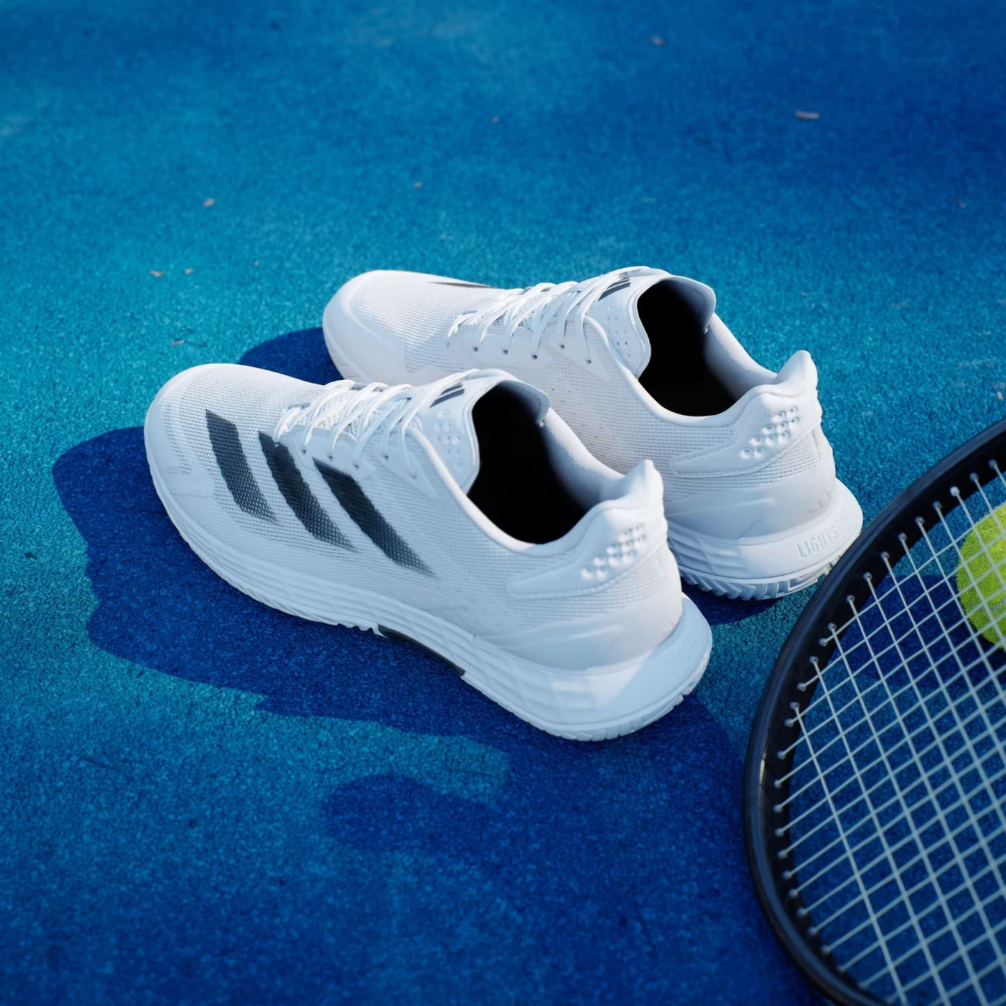 A pair of adidas Defiant Speed 2 Men’s Tennis Shoes in Cloud White/Core Black/Grey sits on a bright blue surface next to a tennis racket with a yellow tennis ball.