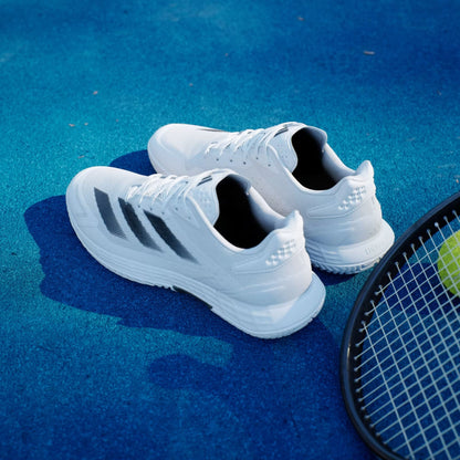 A pair of adidas Defiant Speed 2 Men’s Tennis Shoes in Cloud White/Core Black/Grey sits on a bright blue surface next to a tennis racket with a yellow tennis ball.