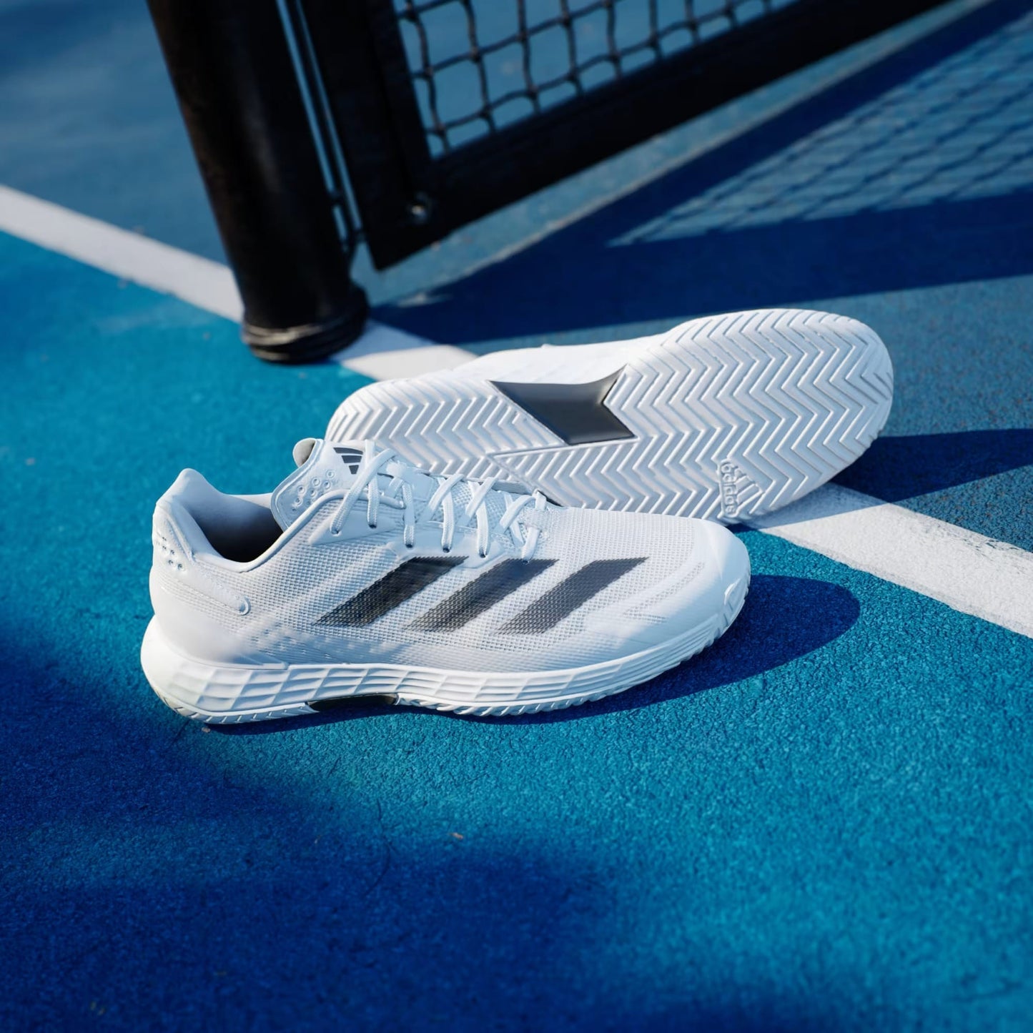 A pair of adidas Defiant Speed 2 Men's Tennis Shoes in Cloud White with Core Black and Grey accents rest on a bright blue outdoor court near a white line and black metal fence.