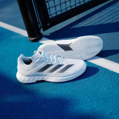 A pair of adidas Defiant Speed 2 Men's Tennis Shoes in Cloud White with Core Black and Grey accents rest on a bright blue outdoor court near a white line and black metal fence.