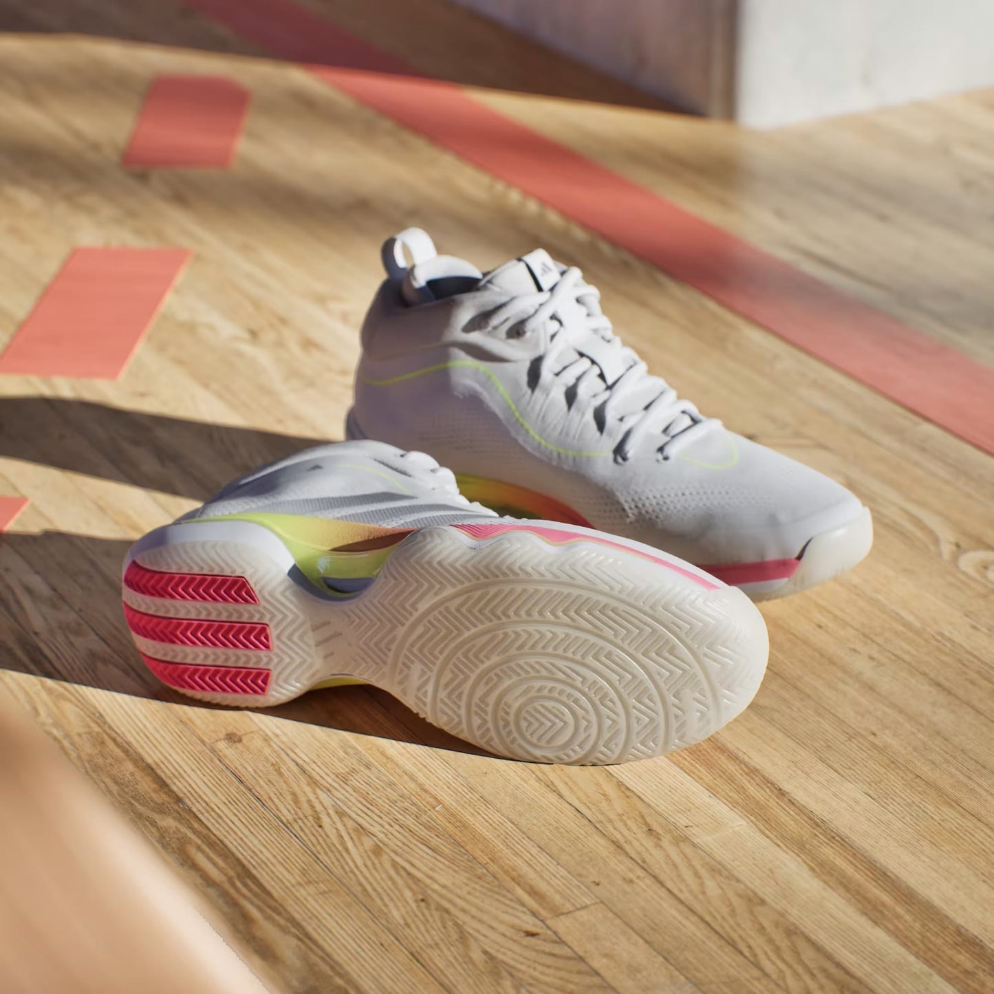 A pair of adidas Crazyflight 7 Men's Indoor Tennis Shoes in Cloud White and Iron Metallic are on a sunlit indoor basketball court—one upright, one on its side showing the textured sole.