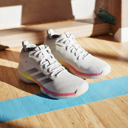 The adidas Crazyflight 7 Mens Indoor Tennis Shoes in Cloud White and Iron Metallic sit on a sunlit wooden floor beside a bright blue line, showcasing their sleek design and comfortable BOOST midsole.