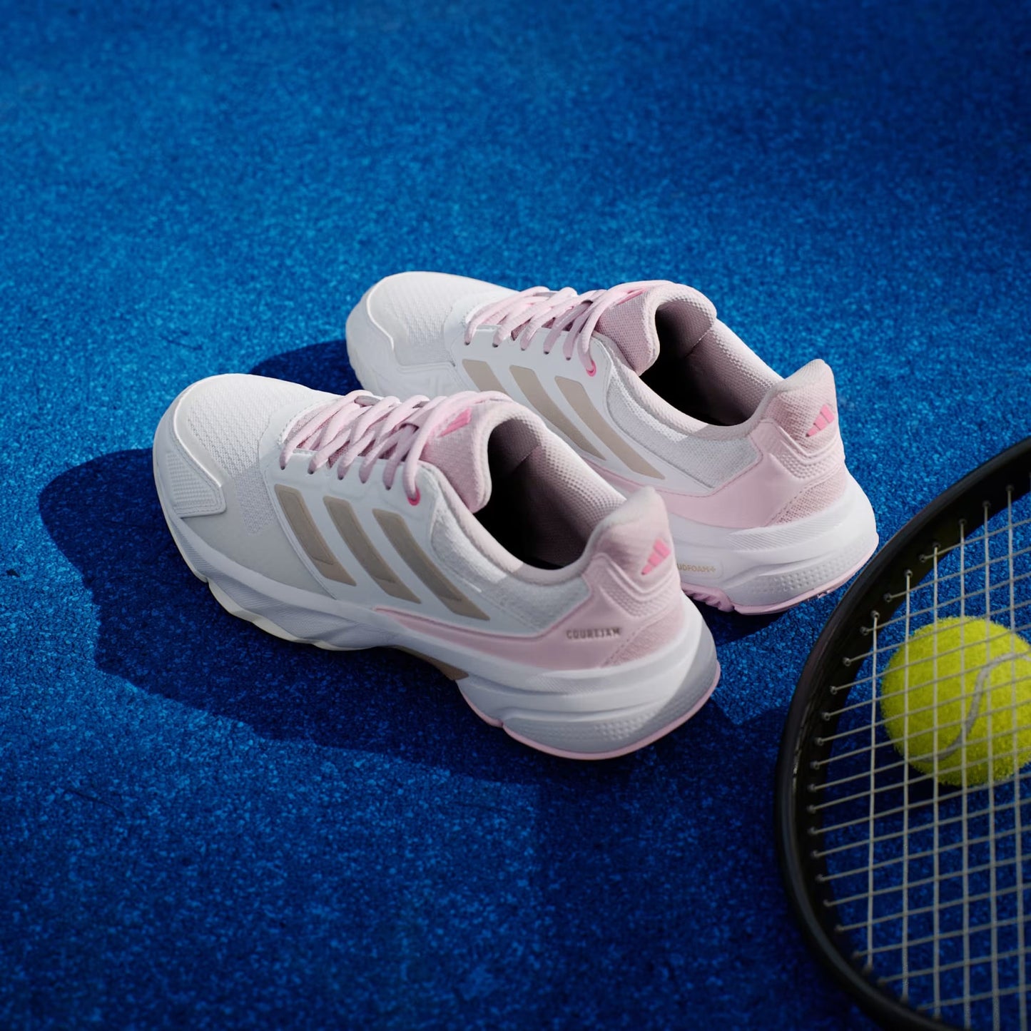 A pair of adidas CourtJam Control 3 Womens Tennis Shoes in Cloud White, Silver Metallic, and Lucid Pink sit on a bright blue hard-court beside a tennis racket and yellow tennis ball.