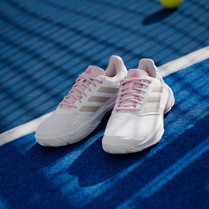 A pair of adidas CourtJam Control 3 Women's Tennis Shoes in Cloud White, Silver Metallic, and Lucid Pink rest on a blue hard court, with a blurred yellow tennis ball and black netting in the background.