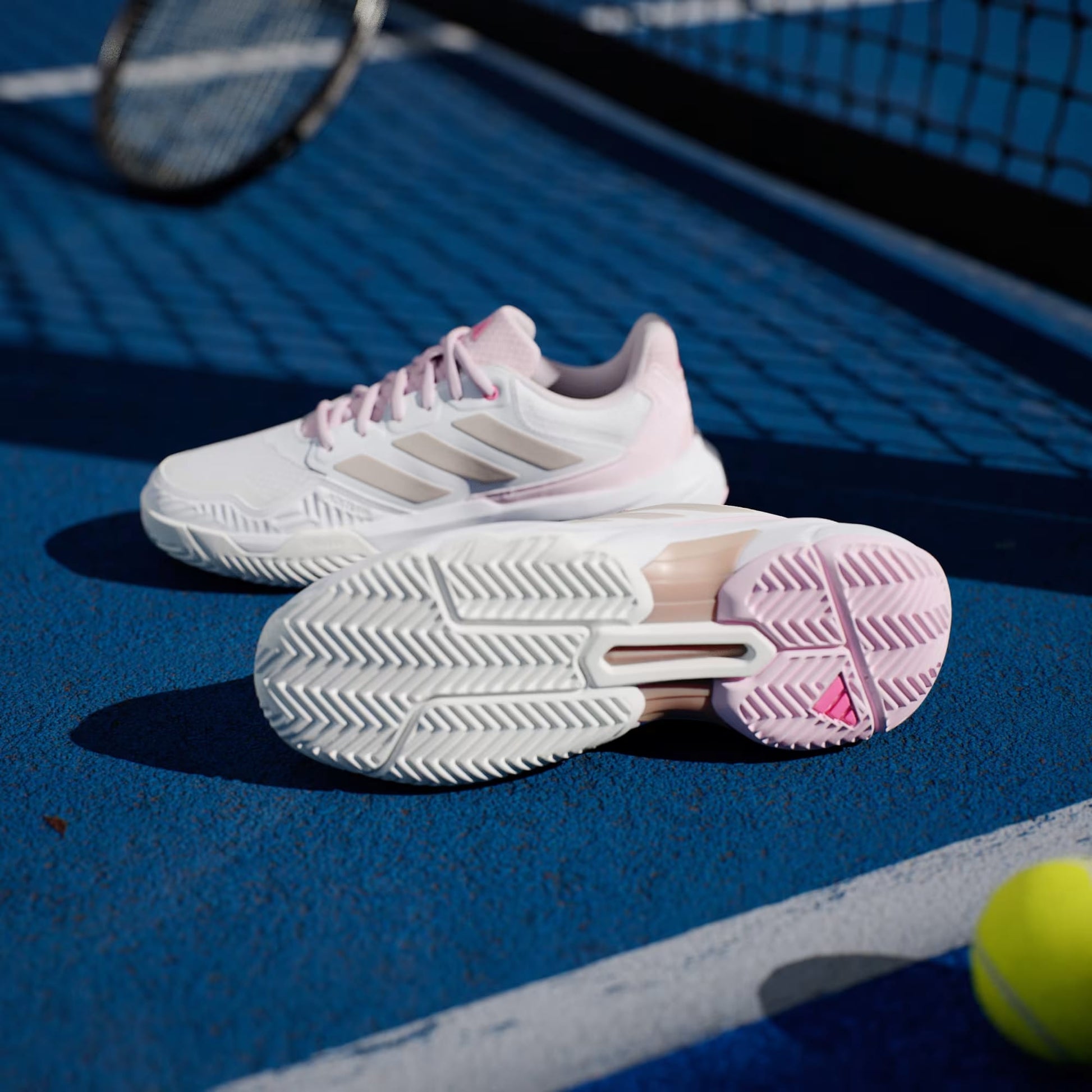 A pair of adidas CourtJam Control 3 Women's Tennis Shoes in Cloud White, Silver Metallic, and Lucid Pink sit on a blue hard court near a net, with a tennis racket and yellow ball in the background.