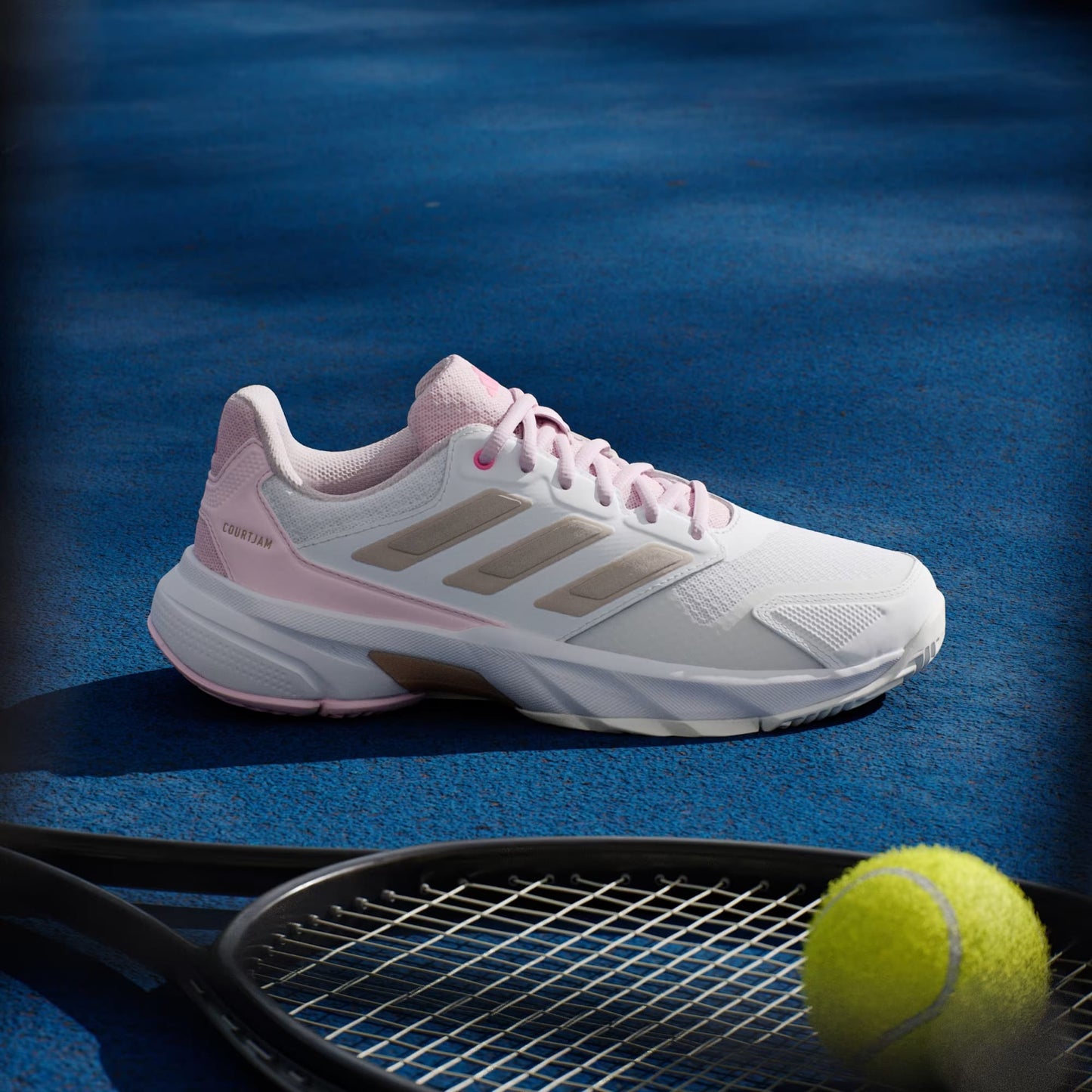 An adidas CourtJam Control 3 Women’s Tennis Shoe in Cloud White, Silver Metallic, and Lucid Pink is displayed on a blue hard court beside a tennis racket and yellow ball.
