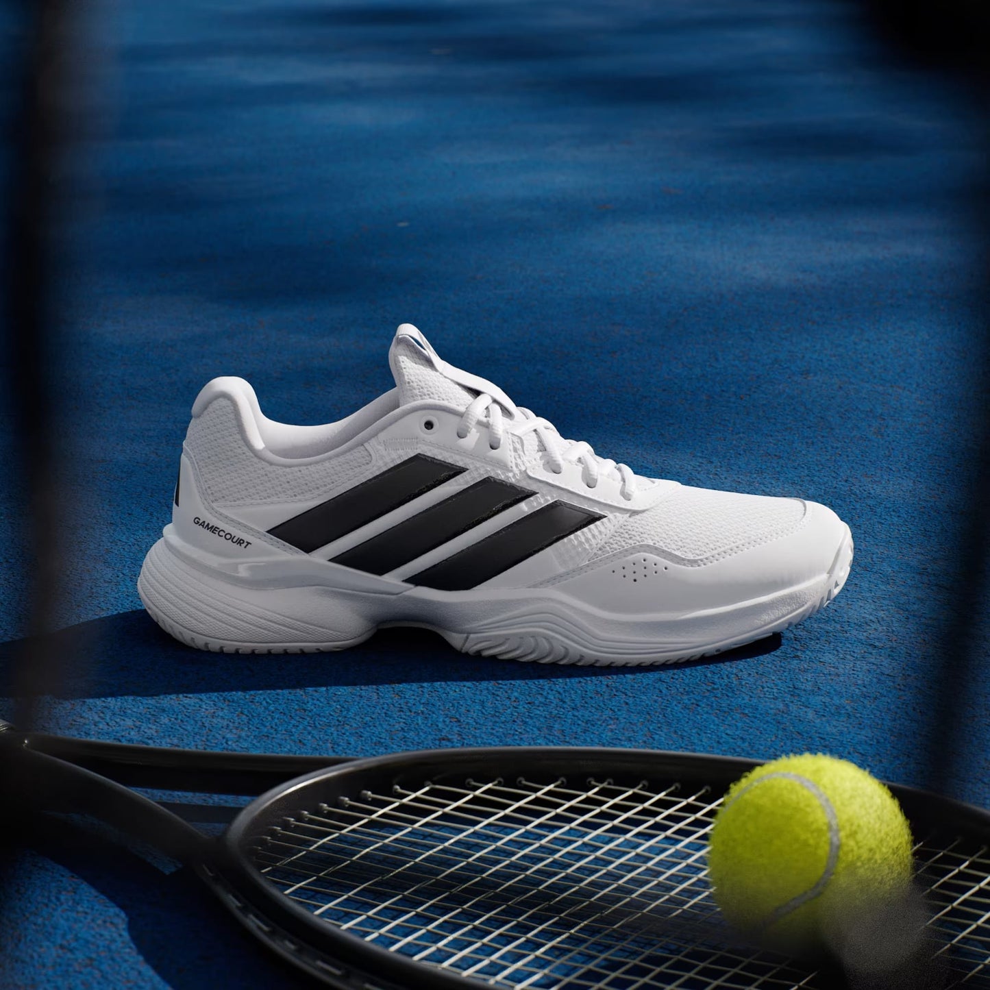 The adidas Gamecourt 3 Men's Tennis Shoes in Cloud White/Core Black sit on a blue court next to a black racket and yellow tennis ball, with racket shadows cast across the lightweight shoe.