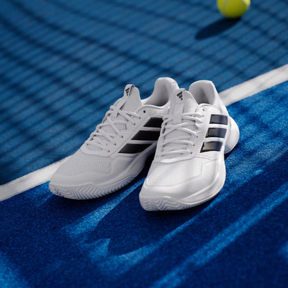 A pair of adidas Gamecourt 3 Mens Tennis Shoes in Cloud White/Core Black sits on a blue tennis court, with a tennis ball and net visible in the background.