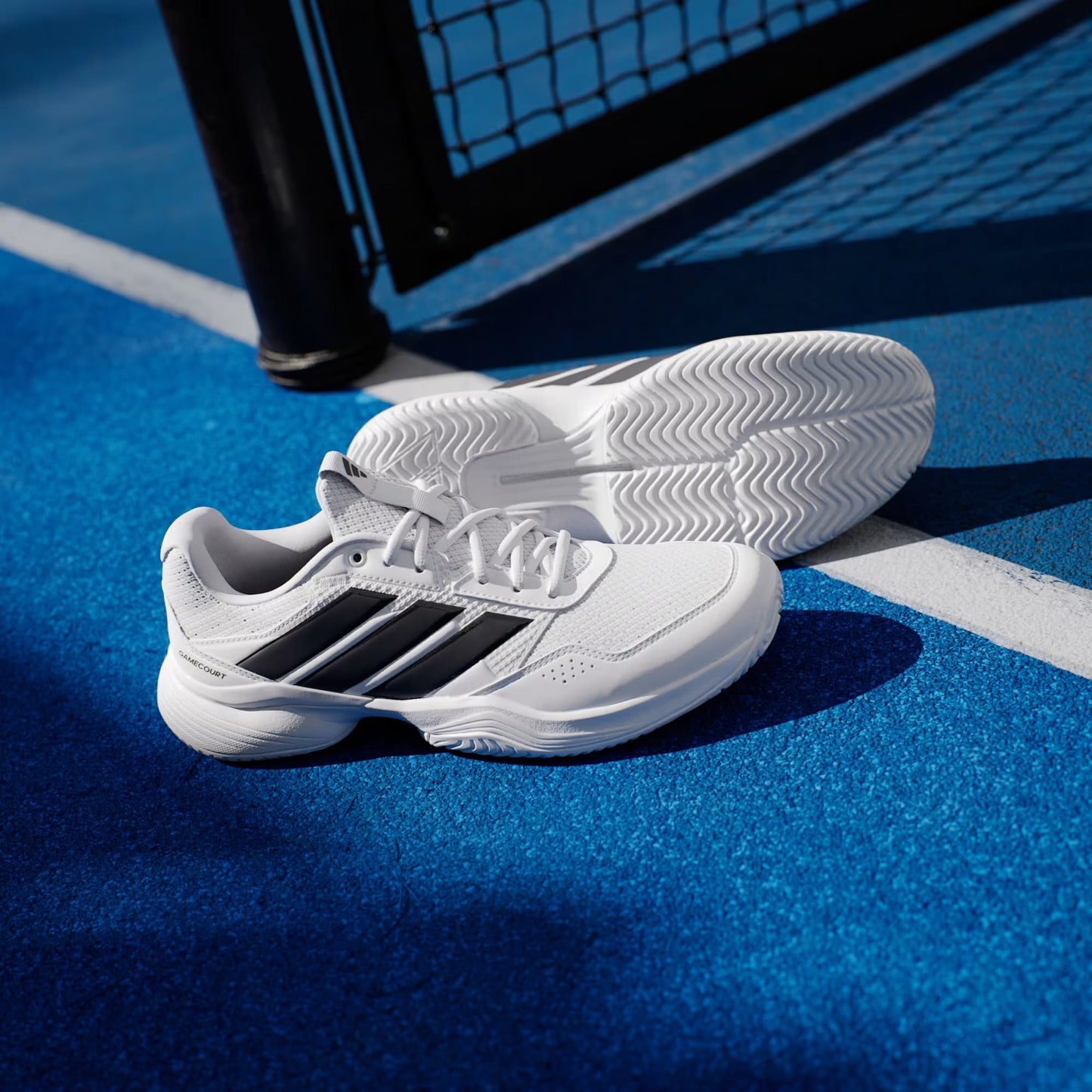 A pair of adidas Gamecourt 3 Men's Tennis Shoes in Cloud White with Core Black stripes sits on a vibrant blue tennis court near a white line and net post.