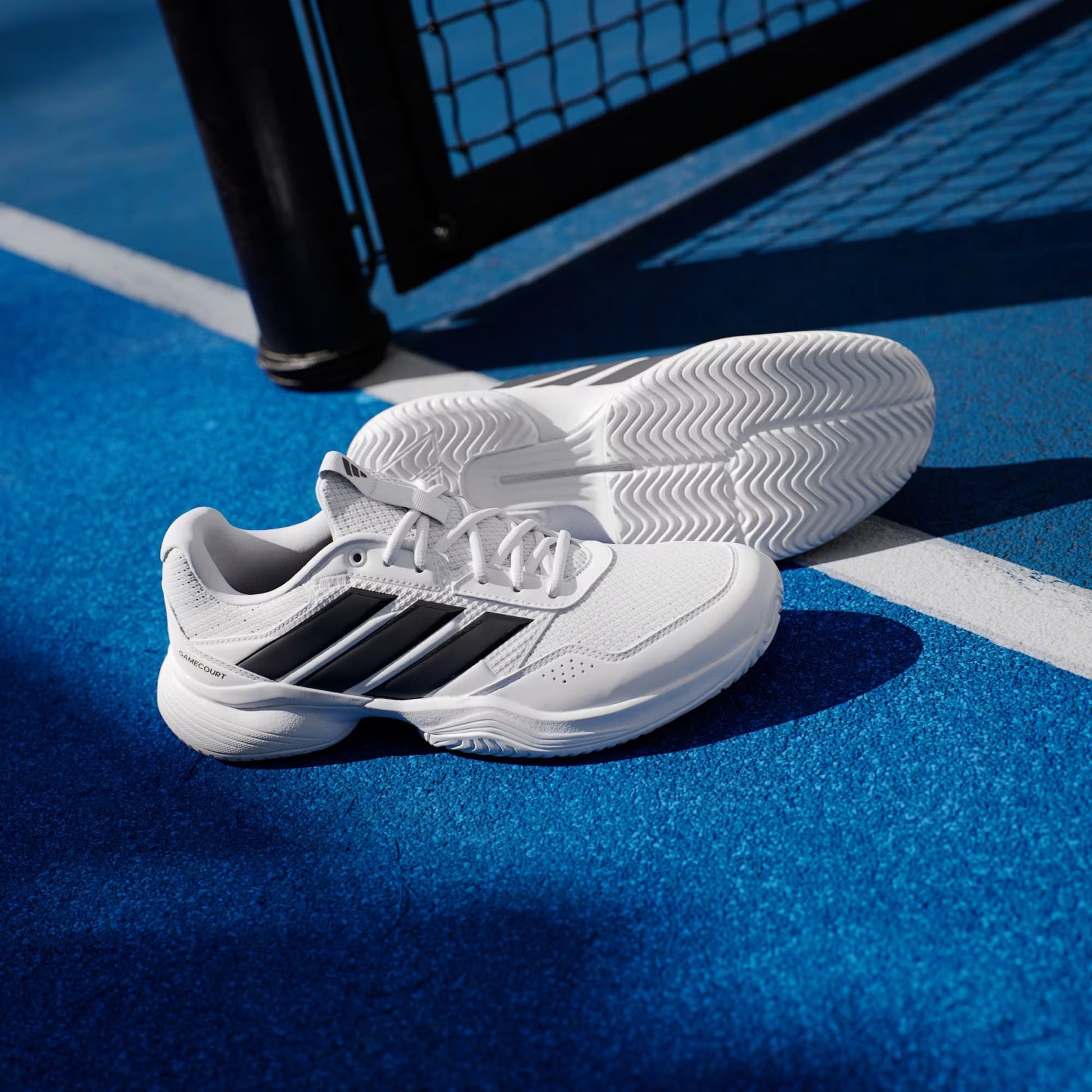 A pair of adidas Gamecourt 3 Men's Tennis Shoes in Cloud White with Core Black stripes sits on a vibrant blue tennis court near a white line and net post.