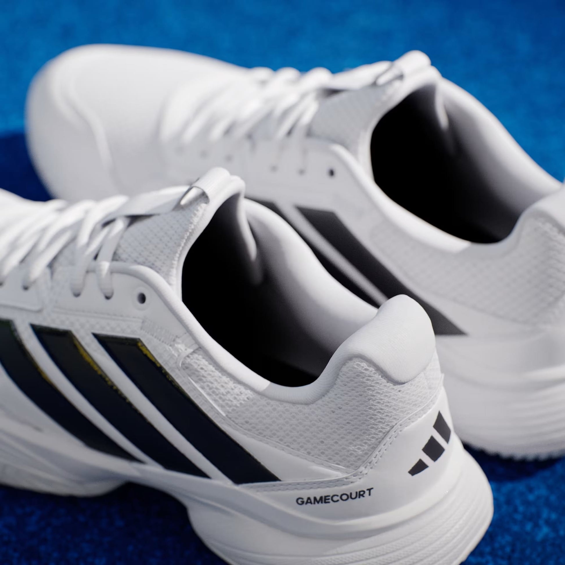 A close-up of adidas Gamecourt 3 Mens Tennis Shoes in Cloud White/Core Black, featuring signature black stripes, placed on a bright blue surface.