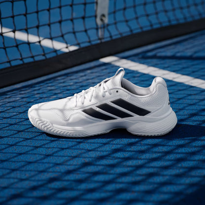 The adidas Gamecourt 3 Men's Tennis Shoes in Cloud White/Core Black rest on a blue court, with black stripes and background shadows from a tennis net.