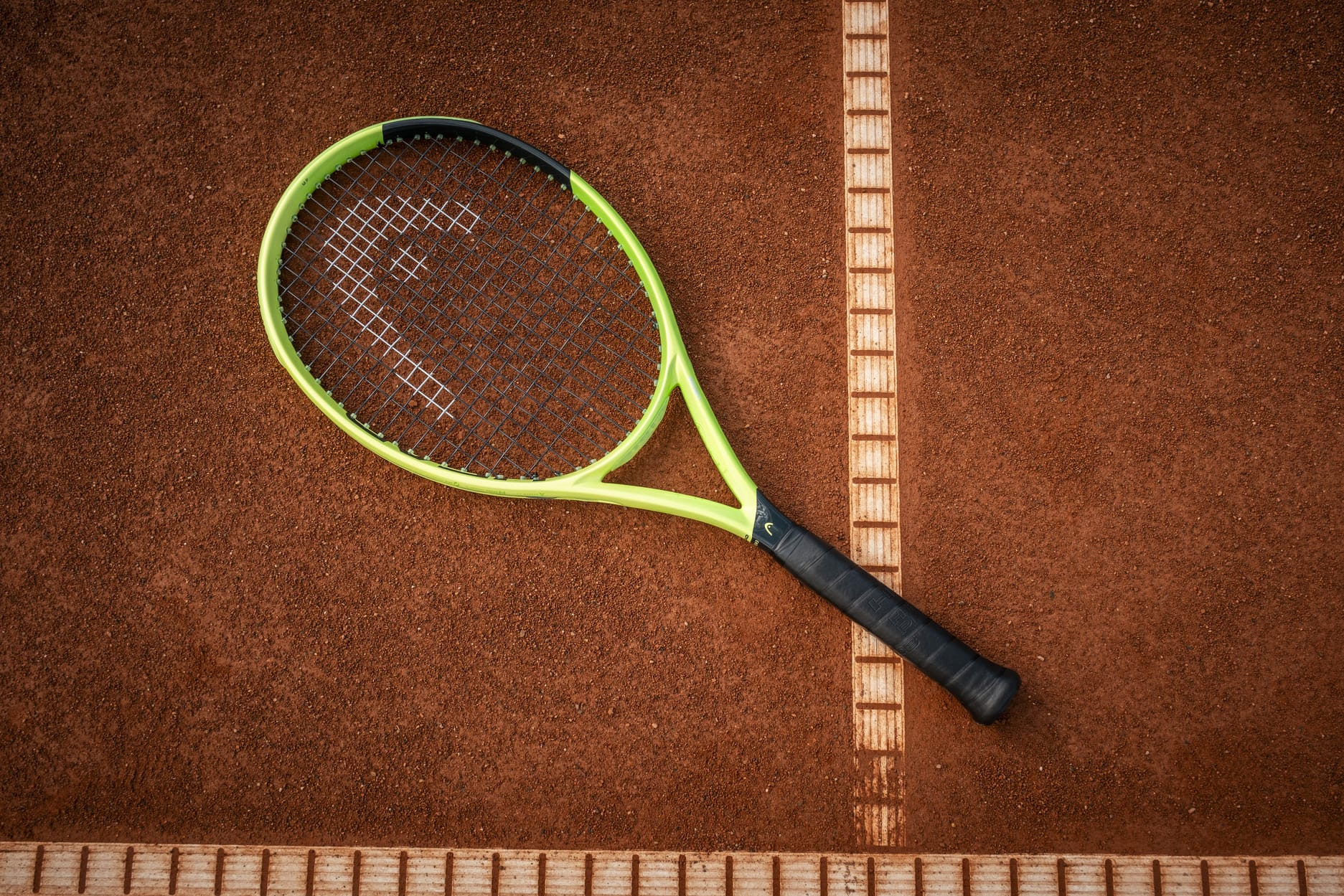 A HEAD Extreme MP 2026 Tennis Racket by HEAD, featuring a green and black design, rests on a clay court with its frame partially over a crisp white boundary line—poised to unleash impressive spin and power with each swing.