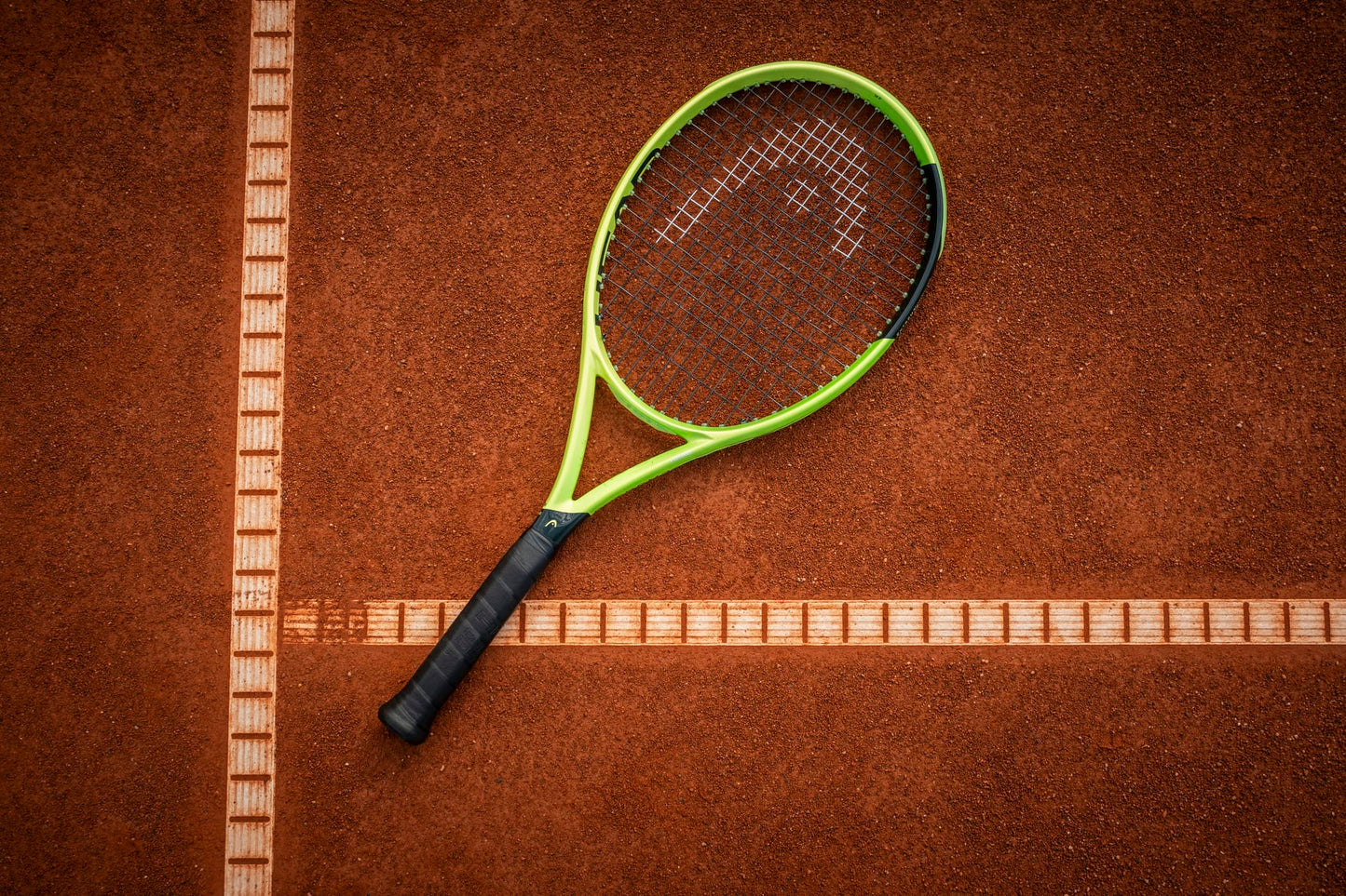 A HEAD Extreme MP 2026 Tennis Racket in striking green and black rests on a clay tennis court, centered at the crossing of two crisp white boundary lines and poised to deliver exceptional spin and power with every swing.