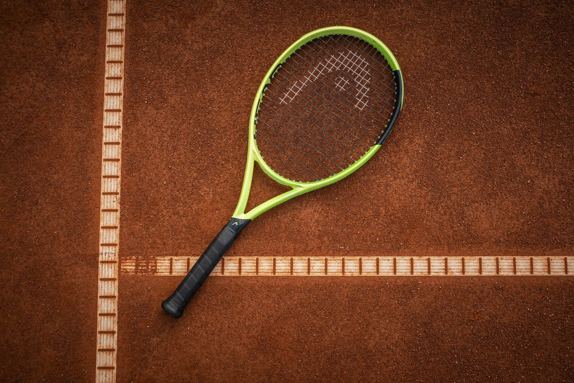 A HEAD Extreme MP 2026 Tennis Racket in striking green and black rests on a clay tennis court, centered at the crossing of two crisp white boundary lines and poised to deliver exceptional spin and power with every swing.