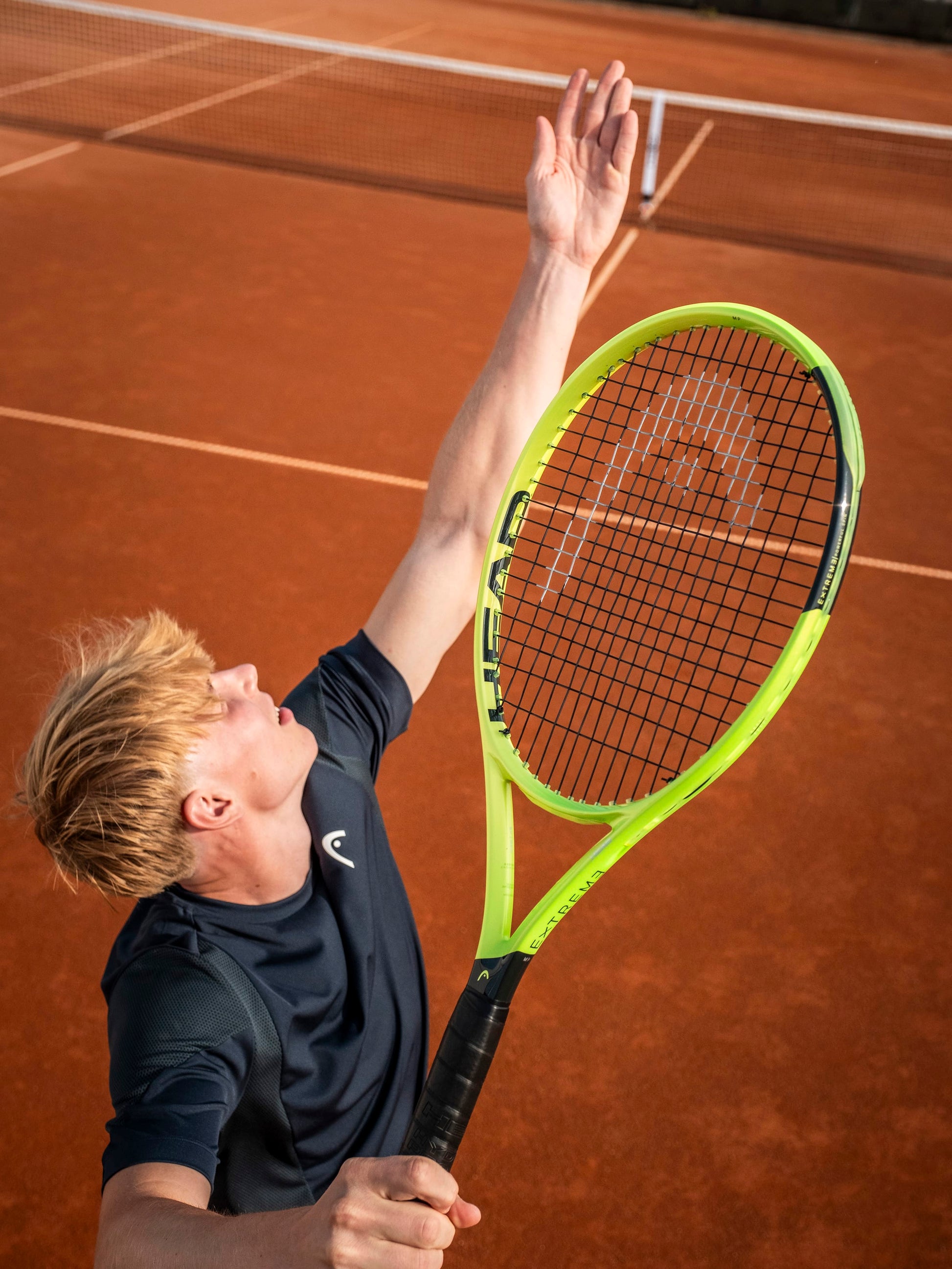 A tennis player wearing a navy shirt prepares to serve on a clay court, tossing the ball while holding the HEAD Extreme MP 2026 Tennis Racket by HEAD, renowned for its impressive spin and power.
