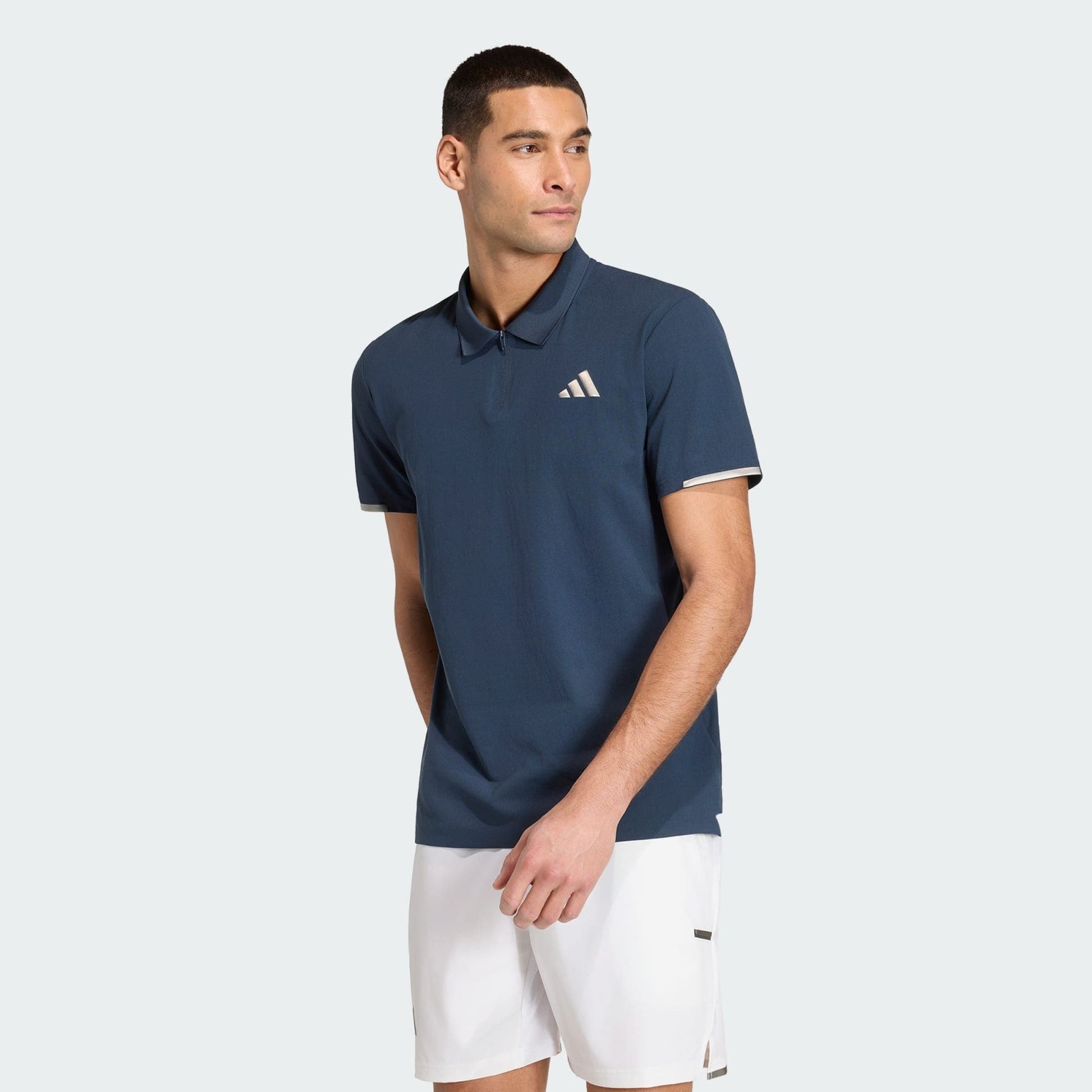 A man in an adidas Climacool Men's Tennis Polo Shirt in Aurora Ink and white shorts stands against a plain light background, looking left with his arms relaxed at his sides.