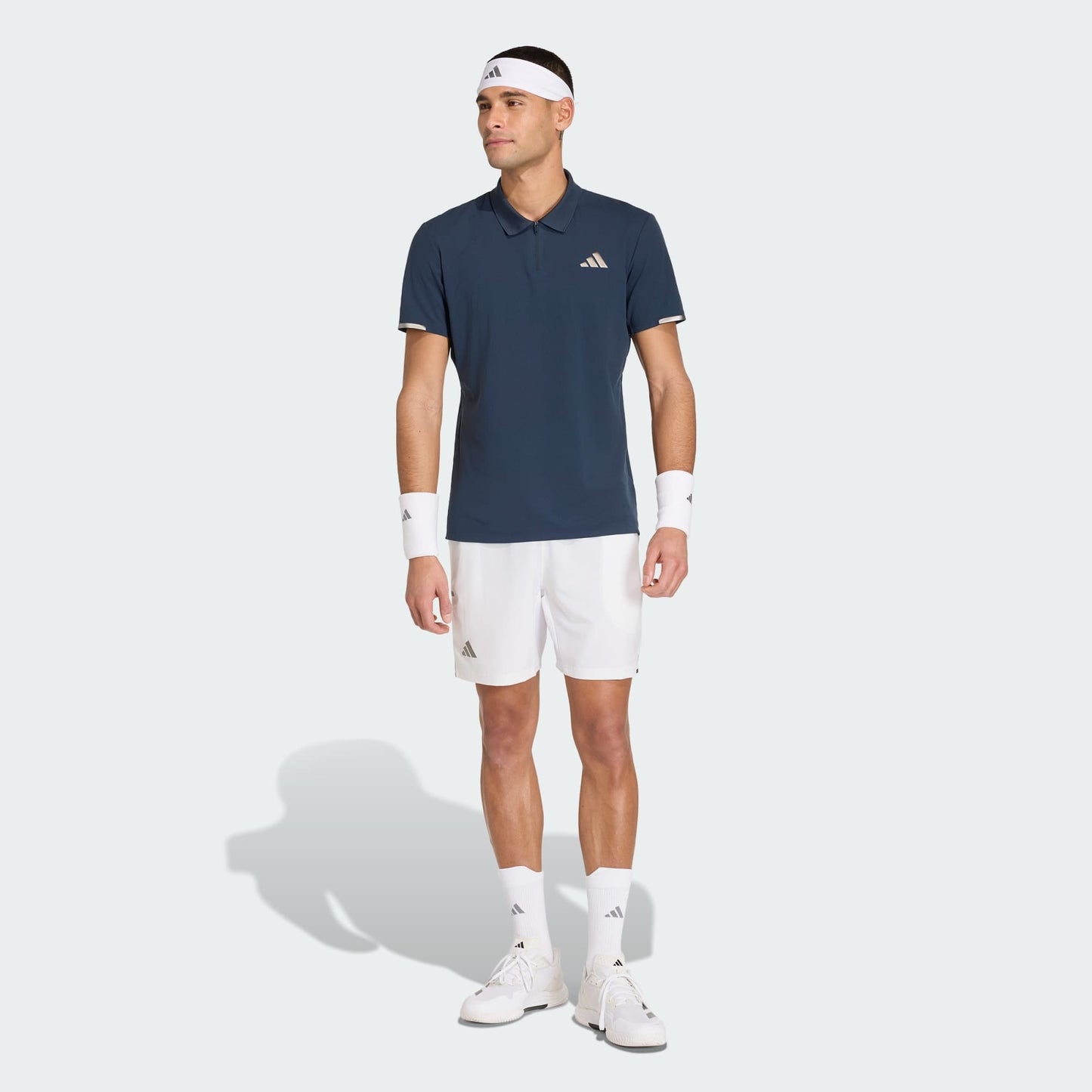 A man wearing an adidas Climacool Men's Tennis Polo Shirt in Aurora Ink, white shorts, white sneakers, socks, wristbands, and a headband stands against a plain light background.