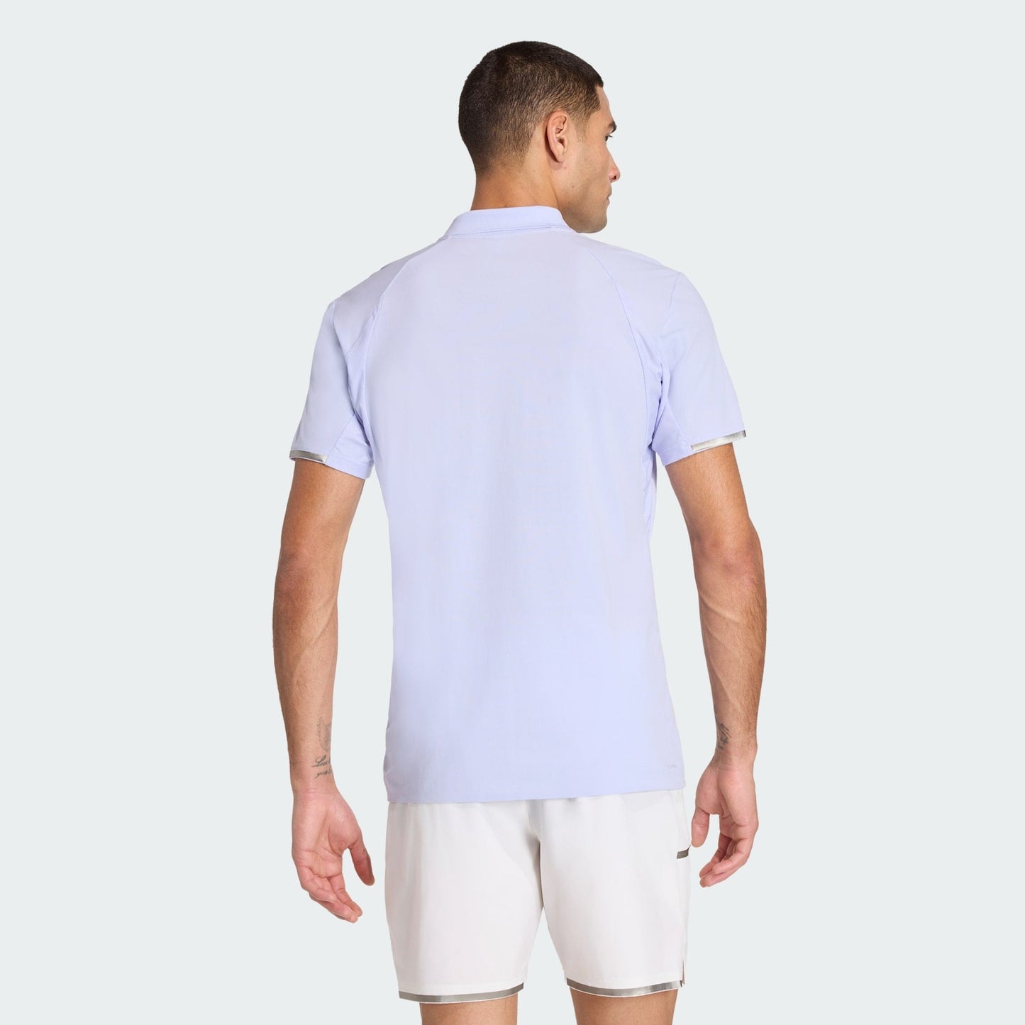 A man faces away from the camera, wearing an adidas Climacool Men's Tennis Polo Shirt in Violet Tone and white shorts, set against a plain light-colored background.