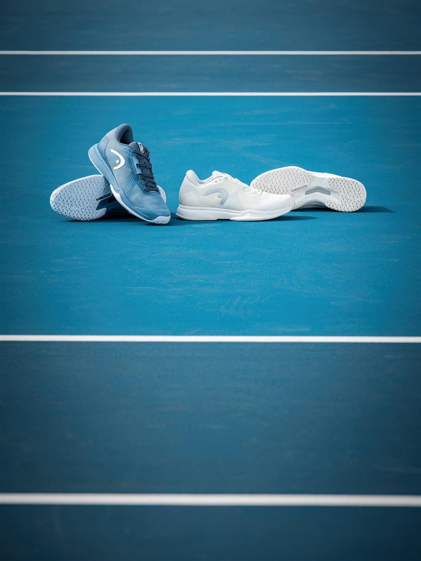 Two pairs of HEAD Sprint Team 4.0 Womens Tennis Shoes by HEAD in Chalk White/Light Grey are displayed side by side on a blue tennis court with visible white lines.