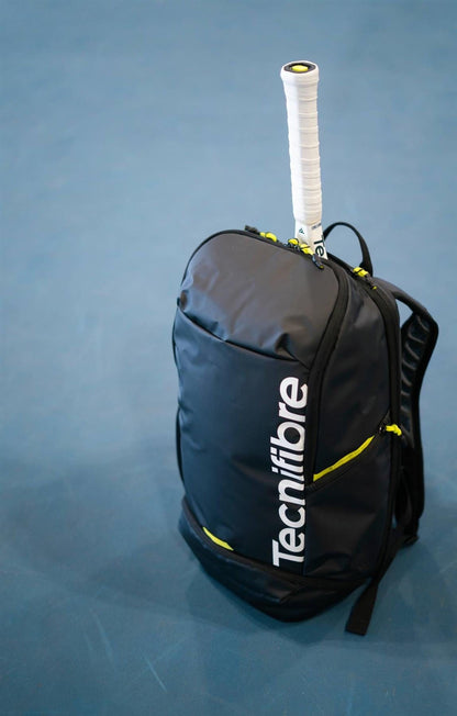 A Tecnifibre Reform Tennis Backpack - Black with a white racket handle sticking out sits on a blue tennis court surface.